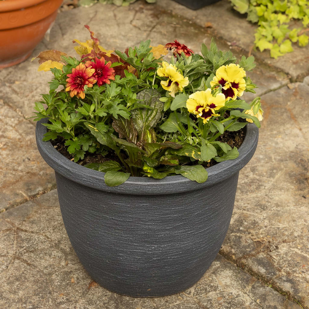 A dark gray textured plastic planter filled with green foliage and colorful flowers, including yellow pansies and red marigolds, set on a stone patio.